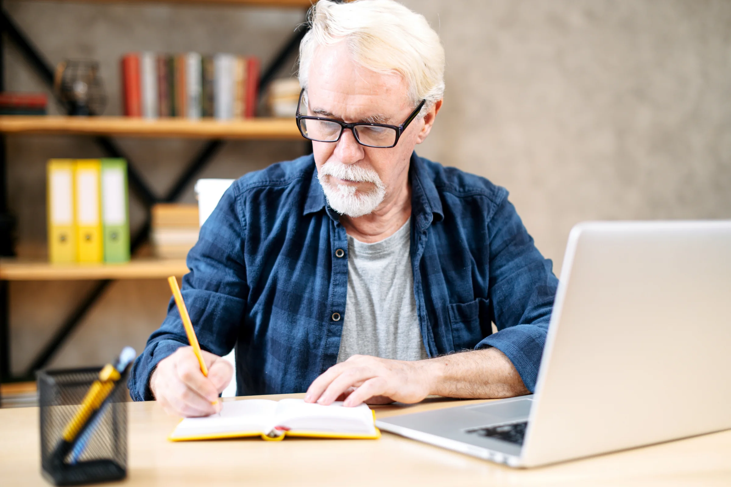 Schmuckbild, älterer Mann am Schreibtisch mit Laptop, Notizbuch und Stift. Er schreibt in das Notizbuch..